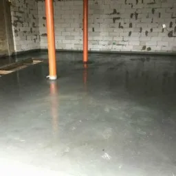 Świeżo wylana, gładka posadzka jastrychowa w surowym wnętrzu, z widocznymi pomarańczowymi rurami pionowymi i fragmentem drewnianego szalunku.