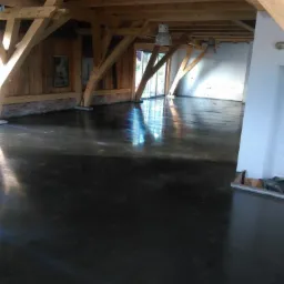 Wnętrze budynku z drewnianą konstrukcją stropu i ciemną, błyszczącą posadzką w trakcie realizacji, widoczne refleksy światła.