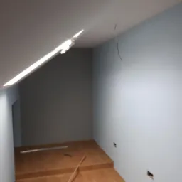 Poddasze po remoncie, ściany pomalowane na jasny niebieski kolor, widoczne przygotowane podłoże pod panele, wystający kabel ze ściany i listwa przypodłogowa na podłodze.