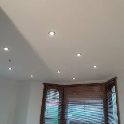 Biały sufit z równomiernie rozmieszczonymi punktowymi lampami LED, widoczne okna z drewnianymi żaluzjami i fragmenty ścian, świeżo po wykończeniu.