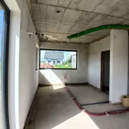 Surowy stan wnętrza z widocznymi przewodami instalacji, sufit betonowy, ściany przygotowane do wykończenia, okno z widokiem na dom.