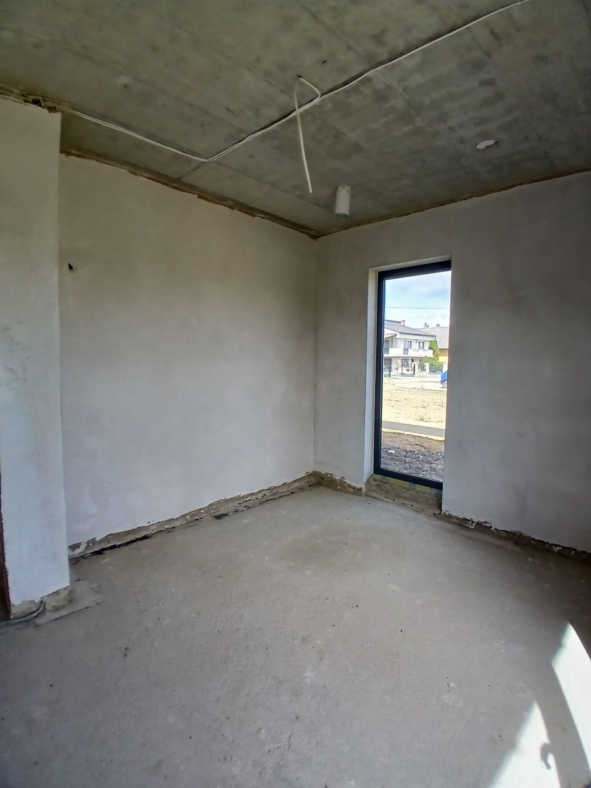 Wnętrze surowego pomieszczenia po nałożeniu tynków na ścianach, widoczne niedoskonałości przy podłodze i surowy betonowy sufit z wystającymi kablami.