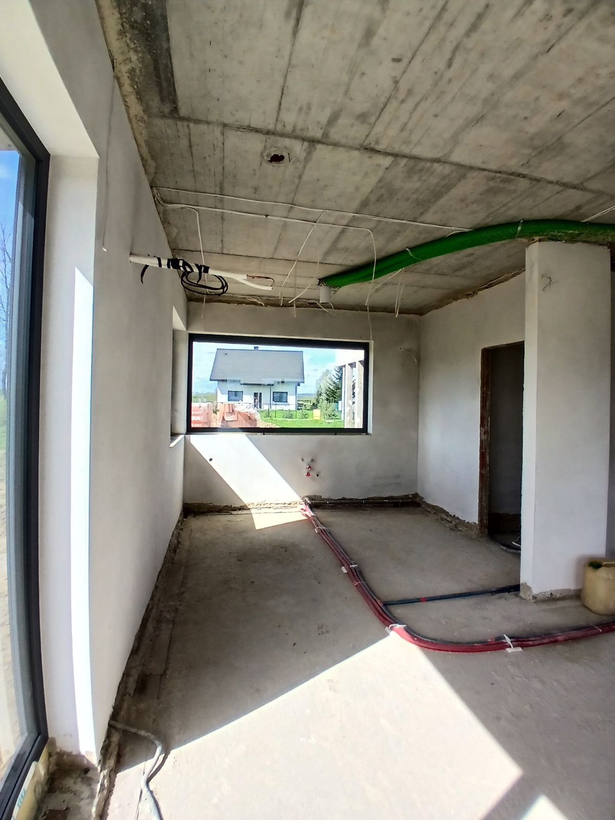 Surowy stan wnętrza z widocznymi przewodami instalacji, sufit betonowy, ściany przygotowane do wykończenia, okno z widokiem na dom.