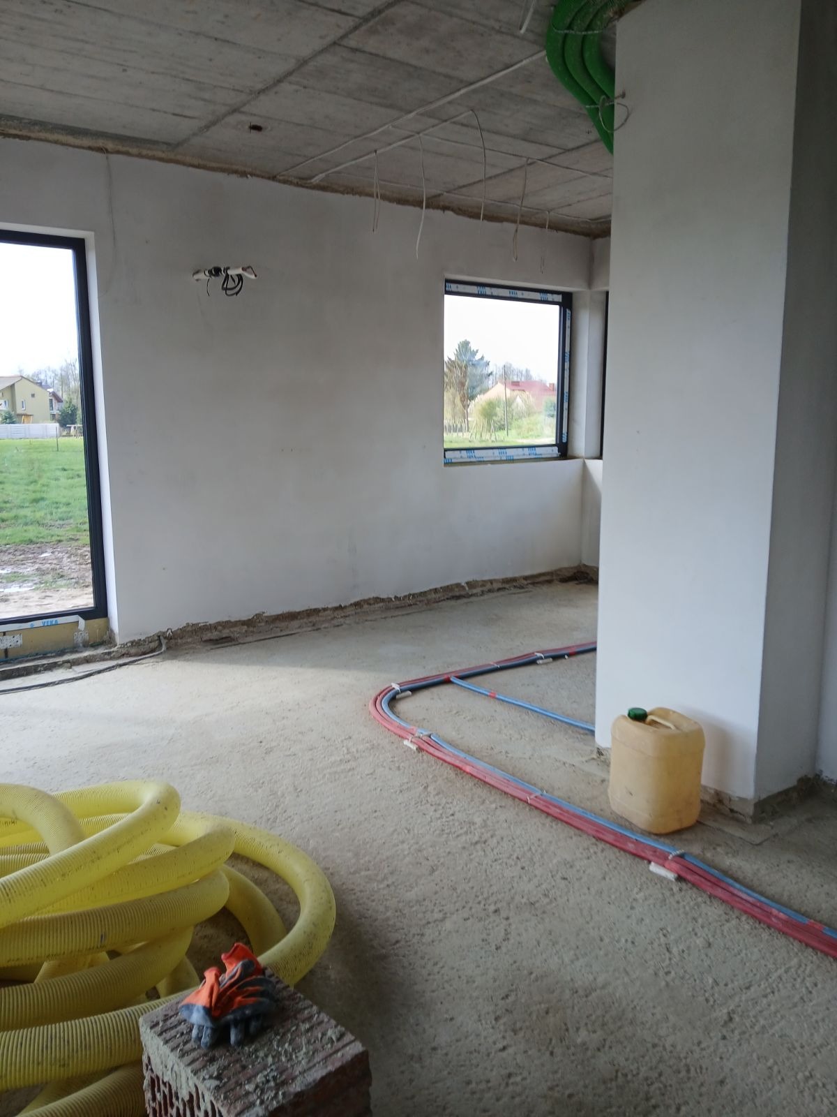 Surowy stan wnętrza budynku w trakcie prac wykończeniowych: widoczne instalacje grzewcze w podłodze, żółte rury wentylacyjne, cegła z rękawicami roboczymi, ściany po wstępnym tynkowaniu.