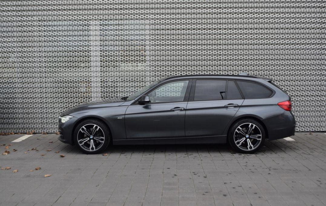 Grafitowy samochód kombi marki BMW zaparkowany na chodniku z szarych płyt na tle metalowej ściany z perforacją.