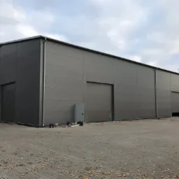 Gotowa hala magazynowa z szarej blachy trapezowej, widoczne trzy bramy wjazdowe, małe drzwi wejściowe, narzędzia na ziemi i jesienne liście na kostce brukowej.