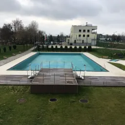 Pusty, prostokątny basen zewnętrzny z drewnianym pomostem w Zbrosławicach, otoczony zielonym trawnikiem i budynkami w tle, sfotografowany przy pochmurnej pogodzie.