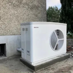 Biała pompa ciepła marki Stiebel Eltron zamontowana przy ścianie budynku z widocznymi rurami odprowadzającymi, na betonowej podstawie.