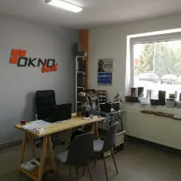 Biuro firmy z ekspozycją profili okiennych przy oknie, logo firmy 'OKNO best' na ścianie, biurko z laptopem i krzesłami.