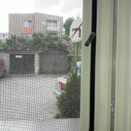 Widok z okna z moskitierą na blok, garaże i znak drogowy zakazu skrętu, uchwyt okienny w pozycji zamkniętej.