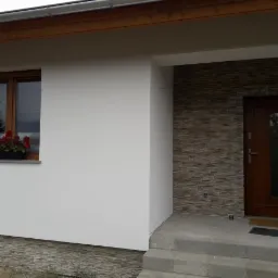 Biała elewacja domu jednorodzinnego w Zielonej Górze z drewnianymi oknami i donicą z czerwonymi kwiatami, oraz wejście z brązowymi drzwiami i dekoracyjnym kamieniem elewacyjnym.