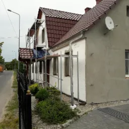 Dom jednorodzinny w trakcie ocieplania, widoczne rusztowania, fragment elewacji z ociepleniem styropianowym i fragment z nałożonym tynkiem, antena satelitarna na ścianie.