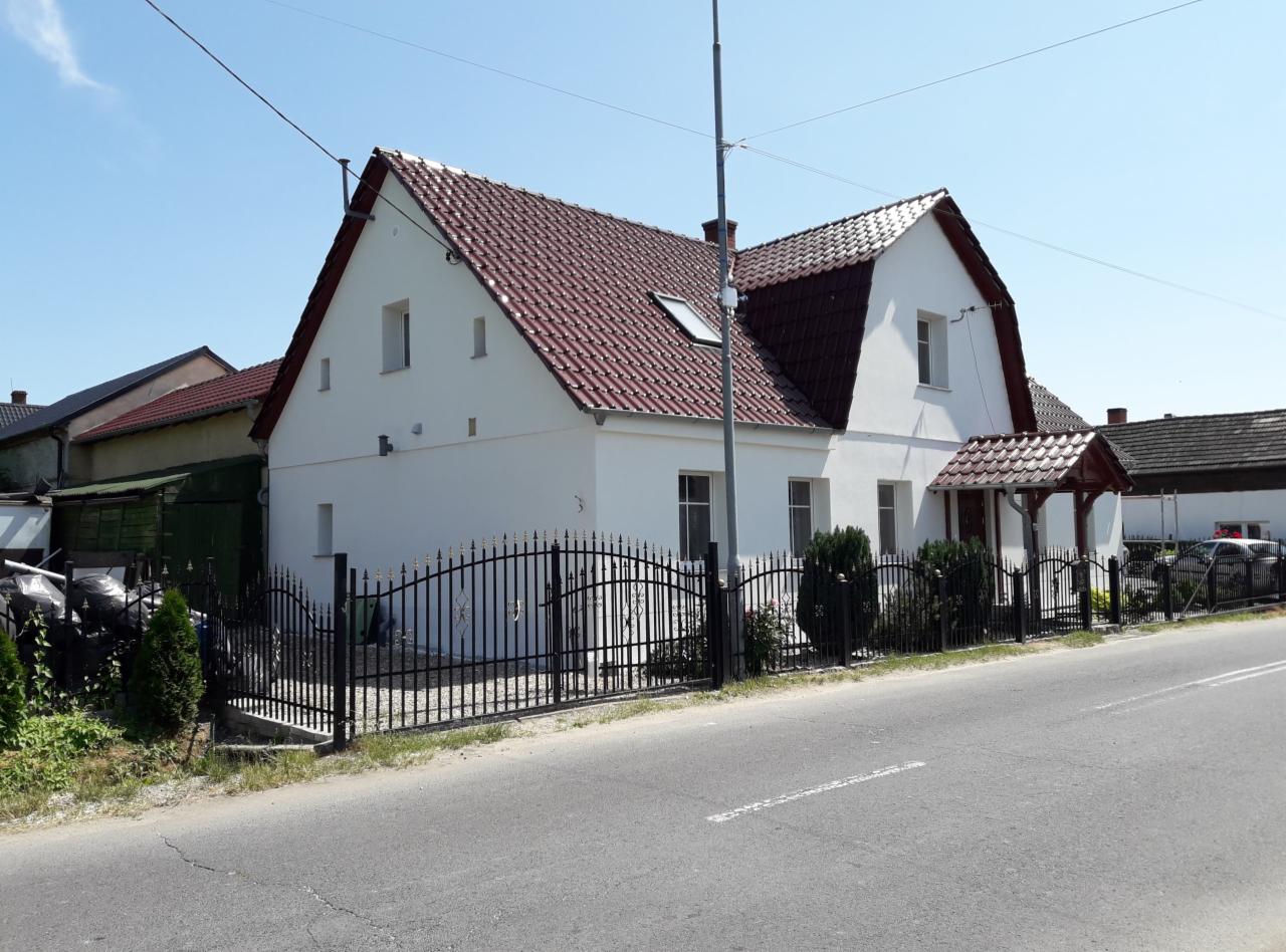 Biały dom jednorodzinny z czerwoną dachówką, czarnym kutym ogrodzeniem z ozdobnymi elementami i niewielkim gankiem z brązową dachówką, widoczny z perspektywy ulicy w słoneczny dzień.