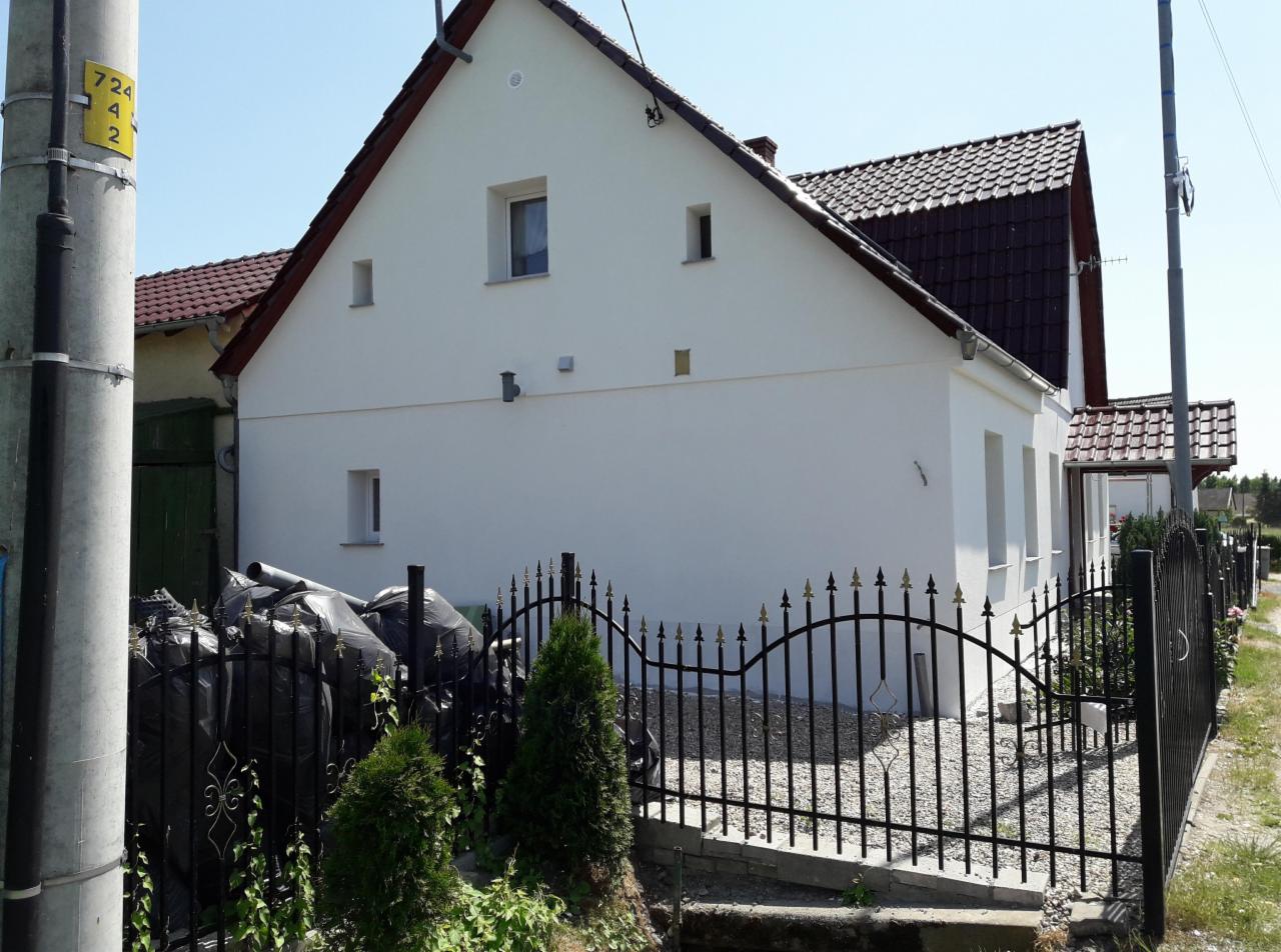 Boczny widok domu po renowacji z białą elewacją, ciemną dachówką i ozdobnym, kutym ogrodzeniem z elementami w kolorze złota, z workami ze śmieciami budowlanymi w tle.