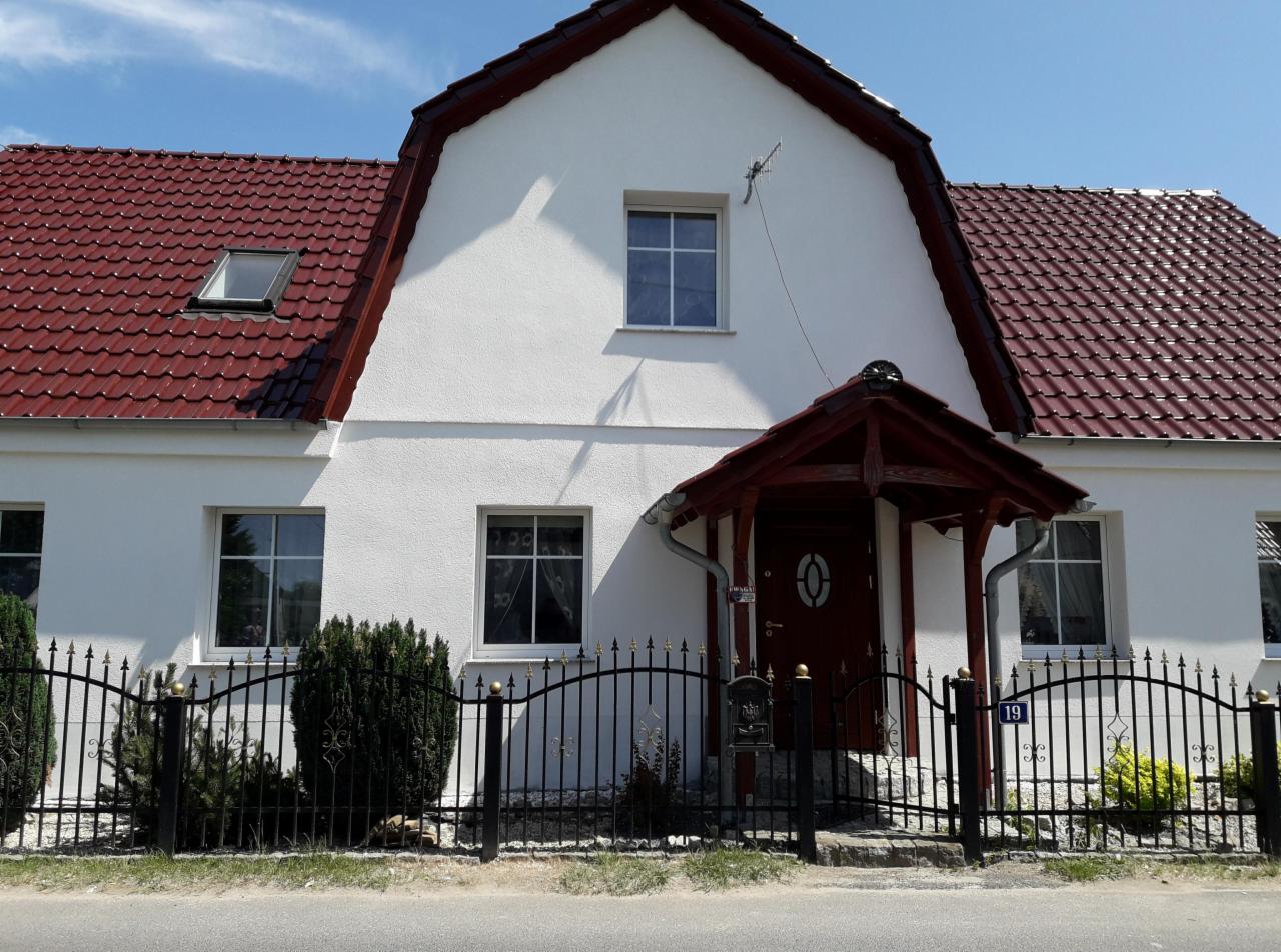 Dom jednorodzinny z białą elewacją, czerwoną dachówką i ozdobnym, kutym ogrodzeniem z numerem 19, widziany z poziomu ulicy w słoneczny dzień.