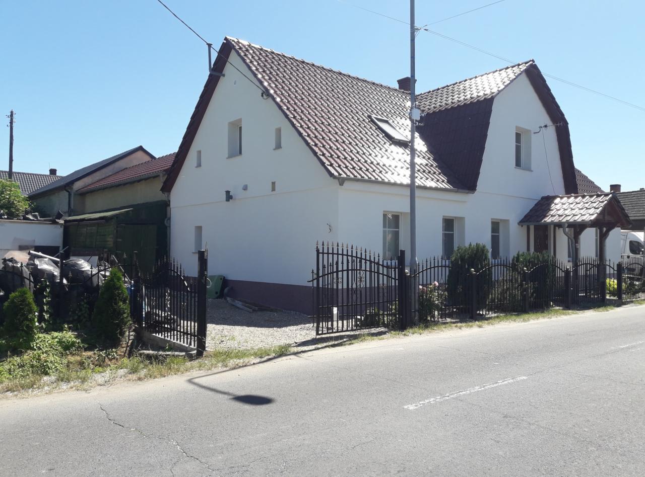 Biały dom jednorodzinny z brązowym dachem, widoczny z poziomu ulicy, z dekoracyjnym, kutym ogrodzeniem i niewielkim podcieniem nad wejściem.