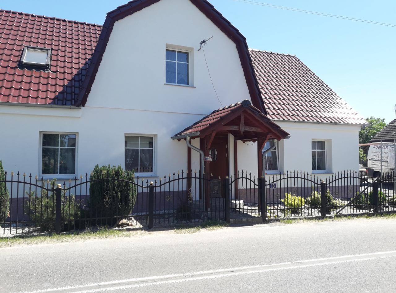 Biały dom jednorodzinny z ciemnoczerwoną dachówką i drewnianą wiatą nad wejściem, widoczny zza ozdobnego, metalowego ogrodzenia. Na posesji zadbana zieleń. W tle zaparkowany samochód dostawczy...