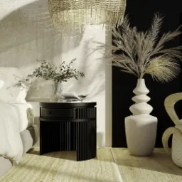 Nowoczesna sypialnia z łóżkiem w jasnych odcieniach, okrągłym czarnym stolikiem nocnym, wazonem z kwiatami i dekoracyjnym oświetleniem wiszącym, uzupełniona o fotel z drewnianą ramą i laptopem w tle.