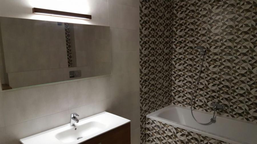 Nowoczesna łazienka z geometrycznymi płytkami w odcieniach brązu i beżu, białą wanną i umywalką z szafką z ciemnego drewna, oświetlona podłużną lampą nad lustrem.