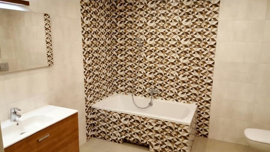 Nowoczesna łazienka z geometrycznymi brązowo-białymi płytkami na ścianie i obudowie wanny, kremowe płytki na pozostałych ścianach, biała wanna i umywalka z szafką z jasnego drewna, lustro...