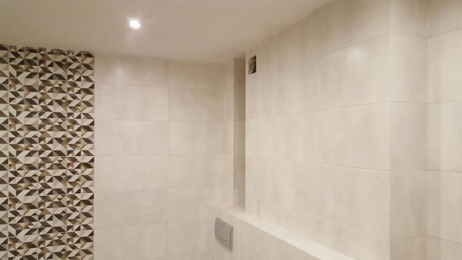 Wykończona łazienka z beżowymi, prostokątnymi płytkami, fragment ściany pokryty mozaiką z geometrycznym wzorem w odcieniach brązu i beżu, widoczny przycisk spłuczki w zabudowie.