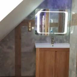 Ukośna łazienka na poddaszu z drewnianą szafką pod umywalką, podświetlanym lustrem, szarą płytką imitującą kamień i brązowymi płytkami podłogowymi.