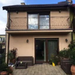 Dwupiętrowy dom z balkonem i tarasem, beżowa elewacja, brązowe balustrady, meble ogrodowe na kostce brukowej, antena satelitarna na dachu.