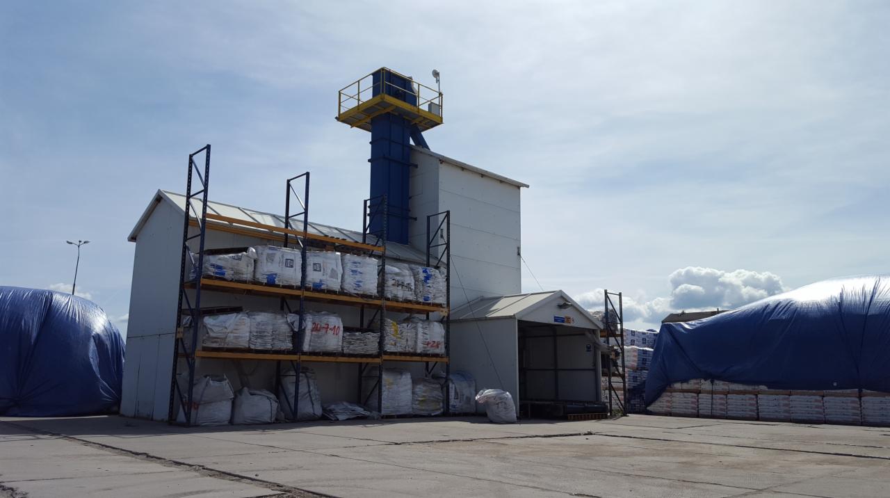 Magazyn zewnętrzny z regałami wypełnionymi workami z materiałami budowlanymi, obok konstrukcja z żółtym pomostem i niebieskim szybem, część worków przykryta niebieską plandeką.