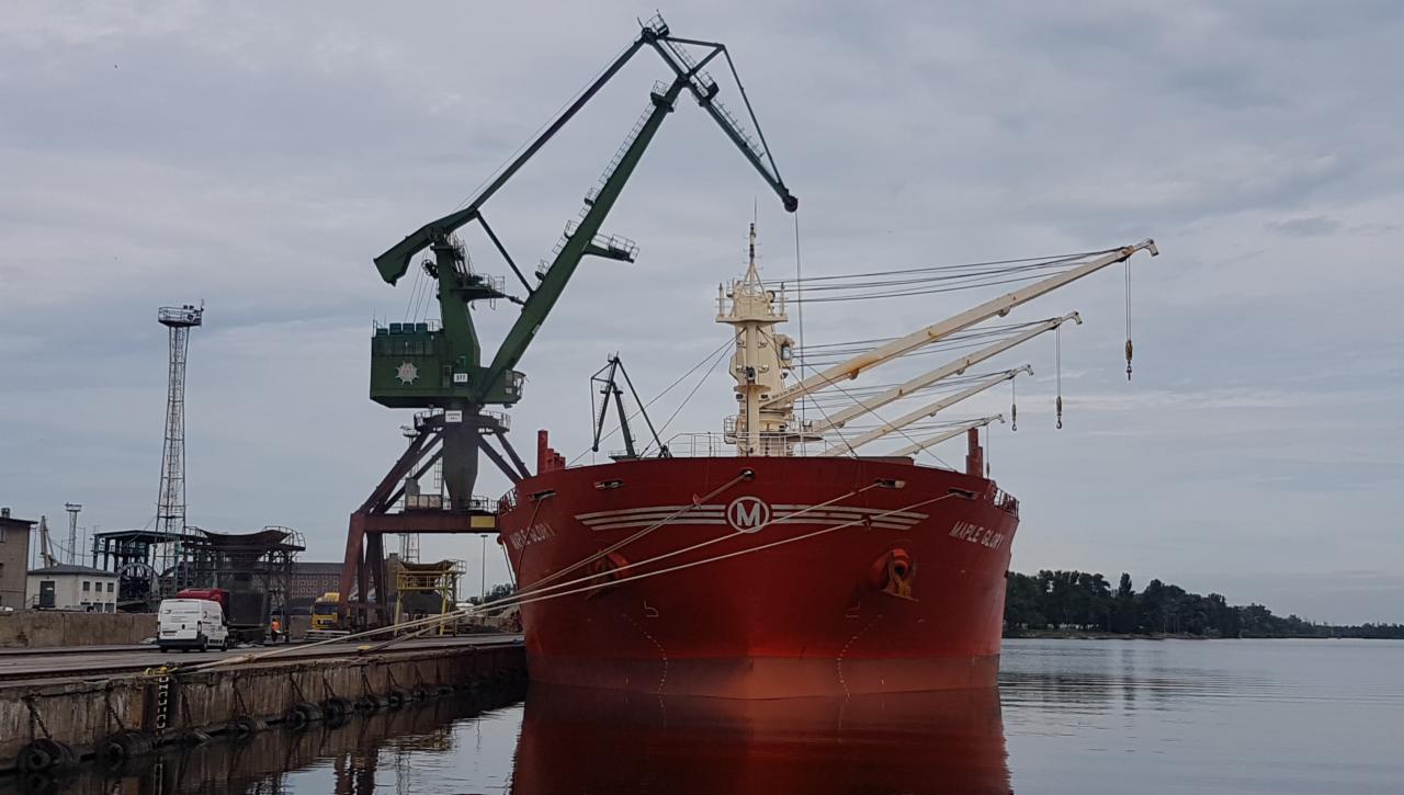 Czerwony statek towarowy Maple Glory cumuje przy nabrzeżu portowym z dźwigami i budynkami w tle, odbijając się w spokojnej wodzie.