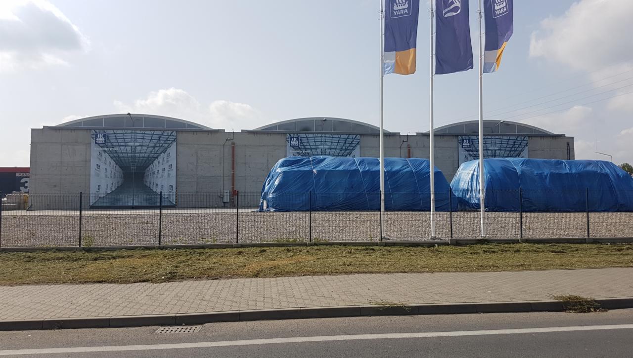 Betonowy magazyn z reklamą w formie perspektywy wnętrza magazynu, z zewnątrz przykryty plandekami, z flagami firmy Yara na maszcie.