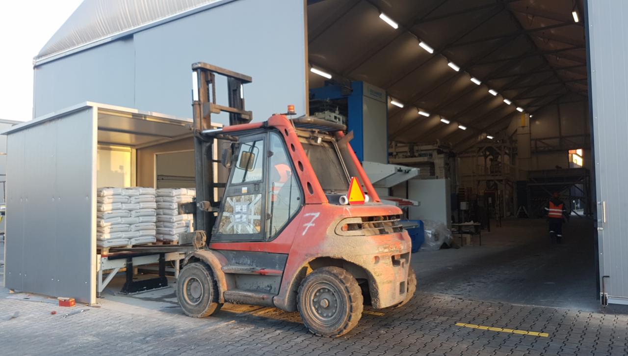 Czerwony wózek widłowy z numerem 7 transportuje paletę z workami w magazynie z konstrukcją stalową i świetlikami dachowymi.