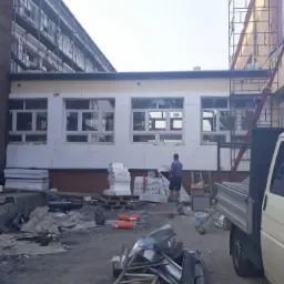 Budynek w trakcie ocieplania, widoczne rusztowania, styropianowe płyty i pracownik w tle, teren budowy z elementami konstrukcyjnymi i samochodem dostawczym.