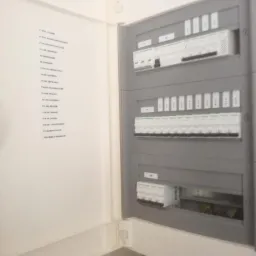 Otwarta szafka rozdzielcza z widocznymi bezpiecznikami i listwą z opisem obwodów na wewnętrznej stronie drzwiczek.