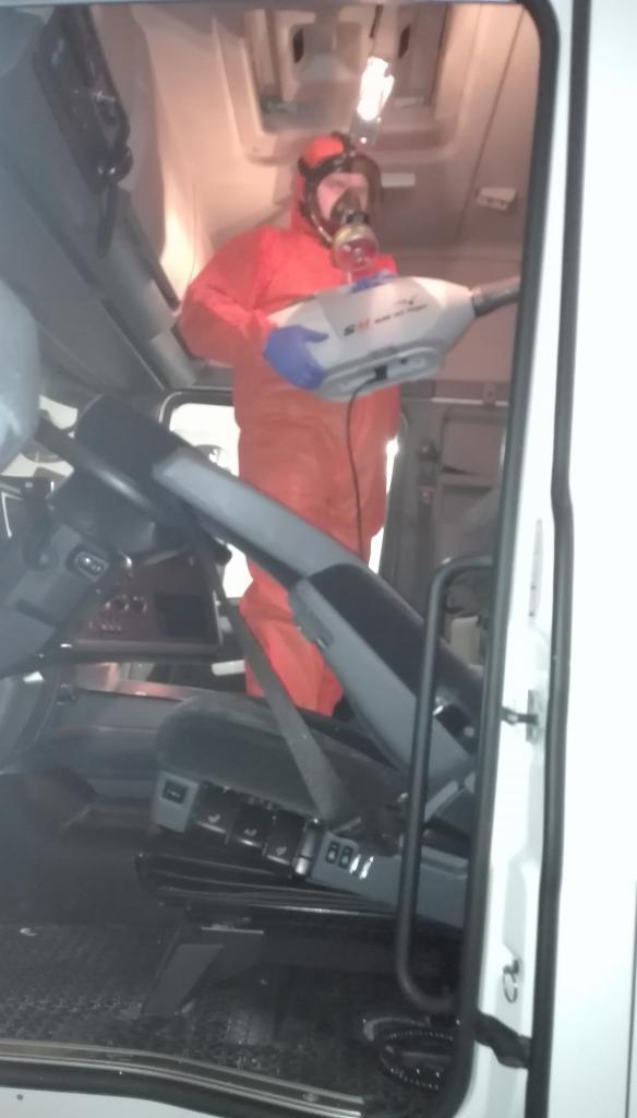 Osoba w kombinezonie ochronnym i masce rozpyla środek dezynfekujący wewnątrz kabiny pojazdu, widoczne elementy kokpitu i fotel kierowcy.