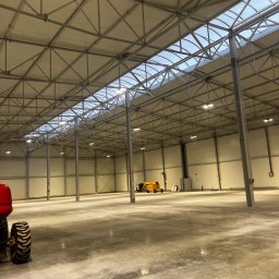 Eektro-Instal lewków - Wnętrze hali magazynowej z widoczną konstrukcją stalową dachu z oknami doświetlającymi, betonową posadzką i częściowo widocznym wózkiem widłowym.