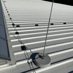 Eektro-Instal lewków - Biały blaszany dach przemysłowy z zainstalowaną, kompletną instalacją odgromową z przewodami i odgromnikiem na betonowej podstawie, widok z góry w słoneczny dzień.