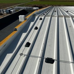 Eektro-Instal lewków - Montaż instalacji odgromowej na dachu z blachy trapezowej, widoczne czarne uchwyty i przewody, w tle krajobraz wiejski.