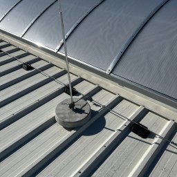 Eektro-Instal lewków - Metalowy dach z zainstalowaną instalacją odgromową: pionowy maszt na betonowej podstawie z przewodami odprowadzającymi, widoczne elementy mocujące i fragment dachu z świetlikiem.