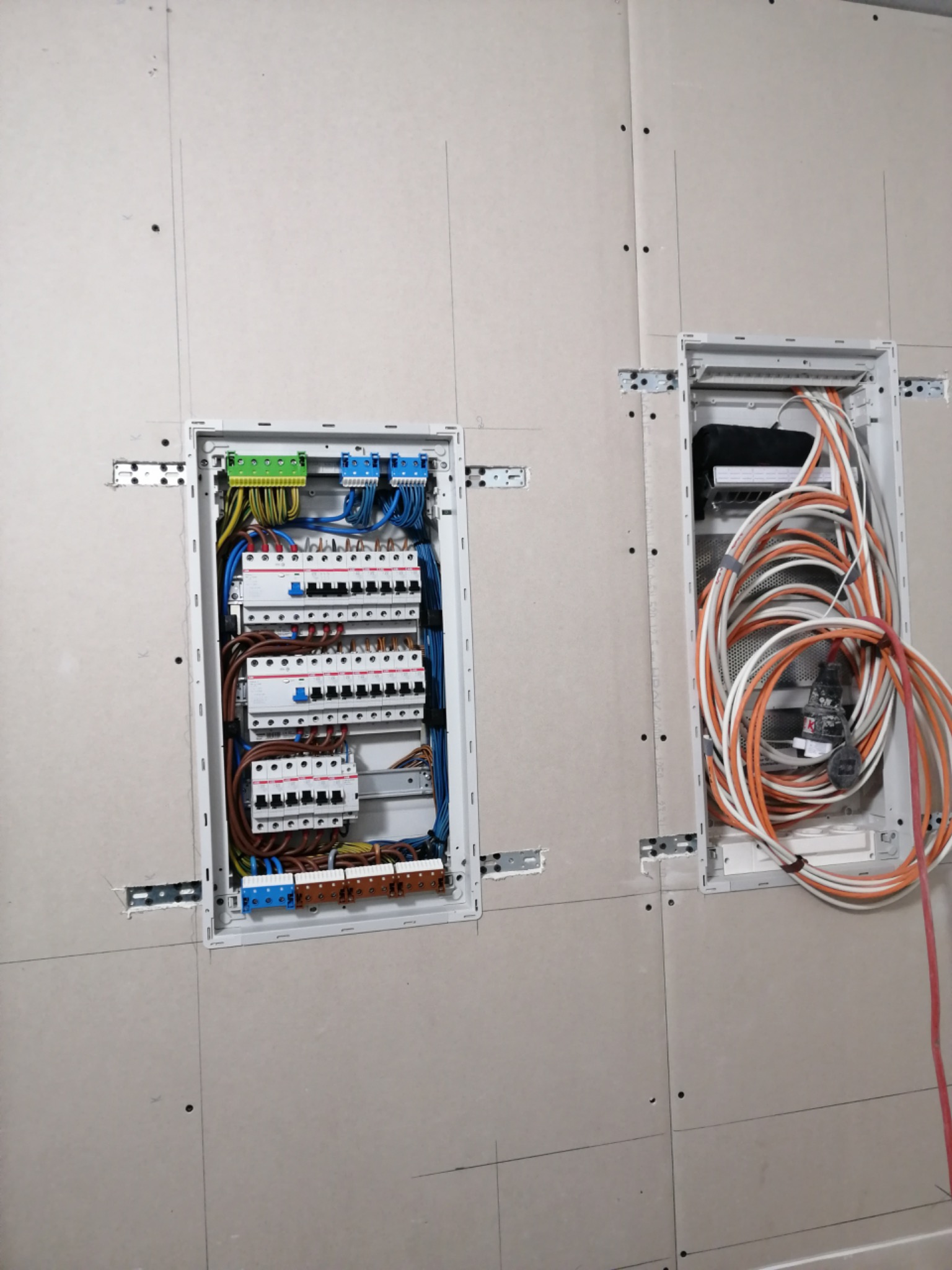 Dwa otwory w surowej ścianie z płyt gipsowych, w jednym zamontowana skrzynka z bezpiecznikami i okablowaniem, w drugim zwój kabli elektrycznych z czarną wtyczką.