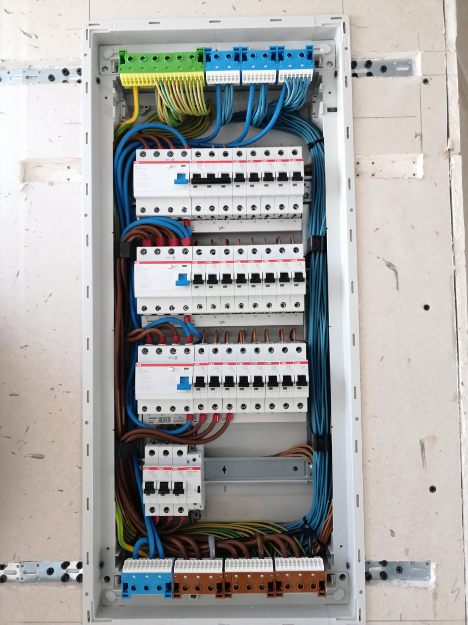 Rozdzielnica elektryczna w obudowie z widocznymi bezpiecznikami, przewodami w kolorach: niebieskim, brązowym, żółto-zielonym, zamontowana w surowej ścianie z widocznymi śladami po montażu.