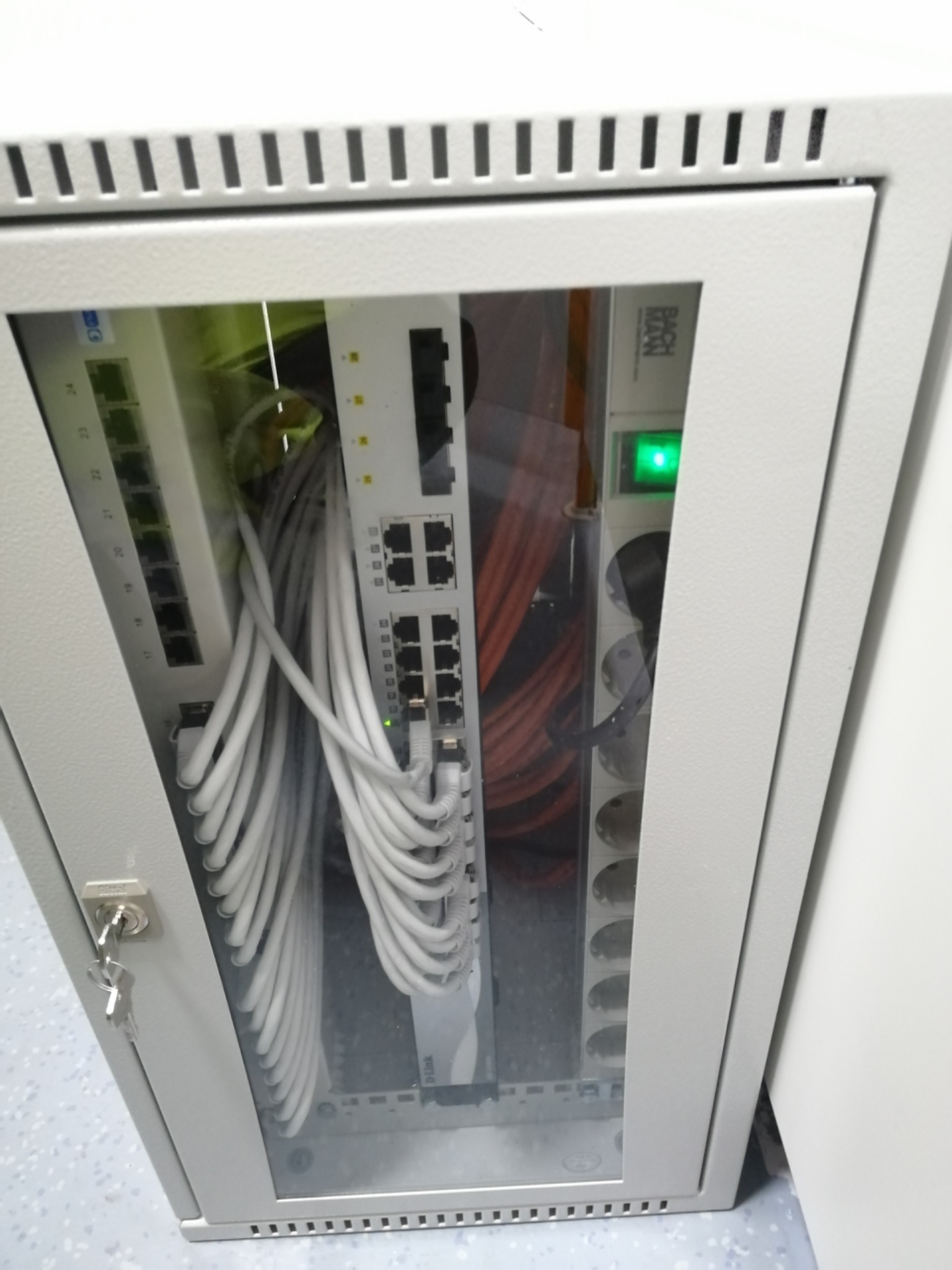Szara szafa rack z widocznymi kablami sieciowymi, przełącznikiem i listwą zasilającą, klucz w zamku drzwi szafy.