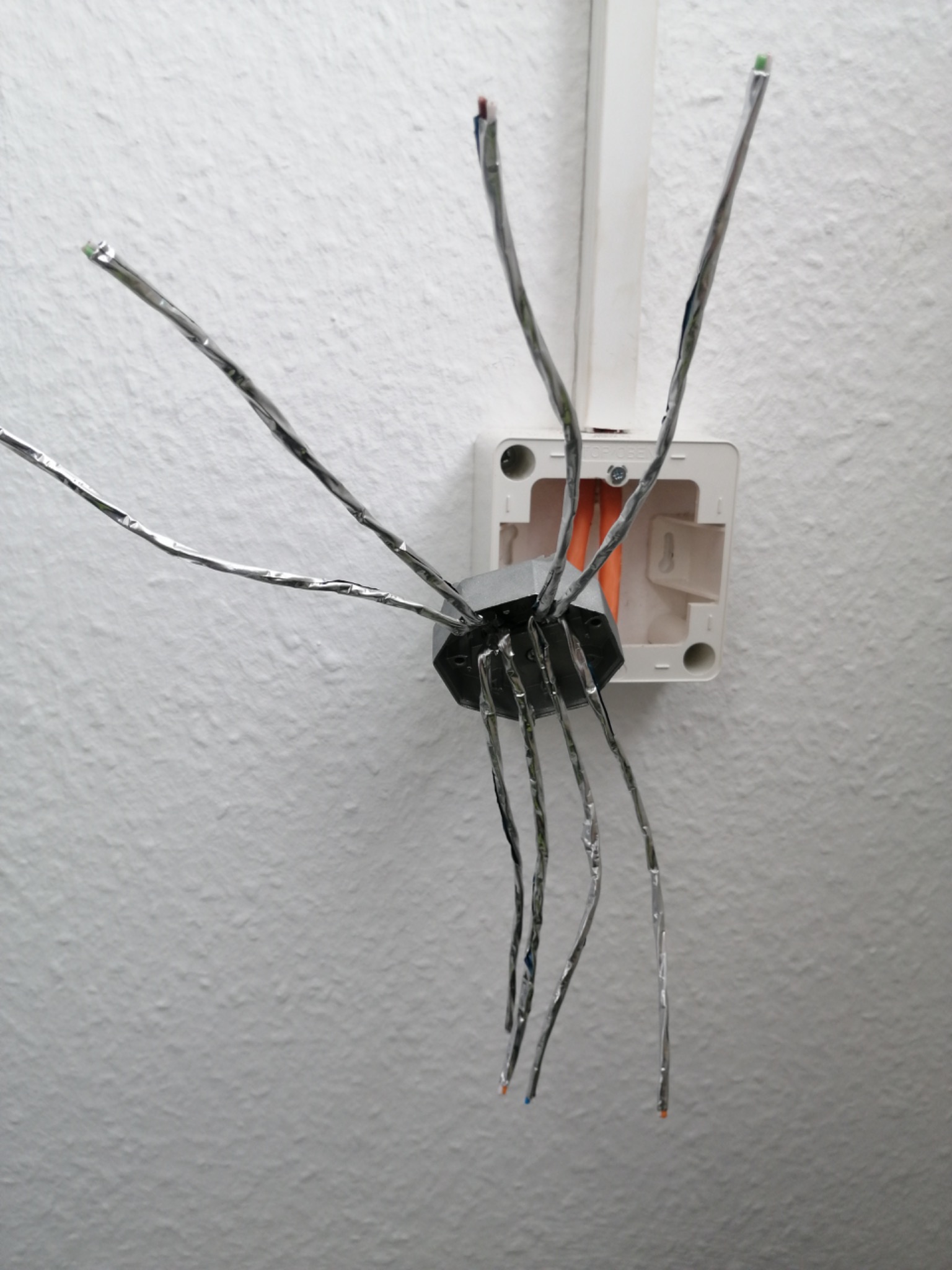 Niezabezpieczone przewody elektryczne owinięte folią aluminiową wystające ze ściany z puszką instalacyjną, widoczne kolory izolacji.