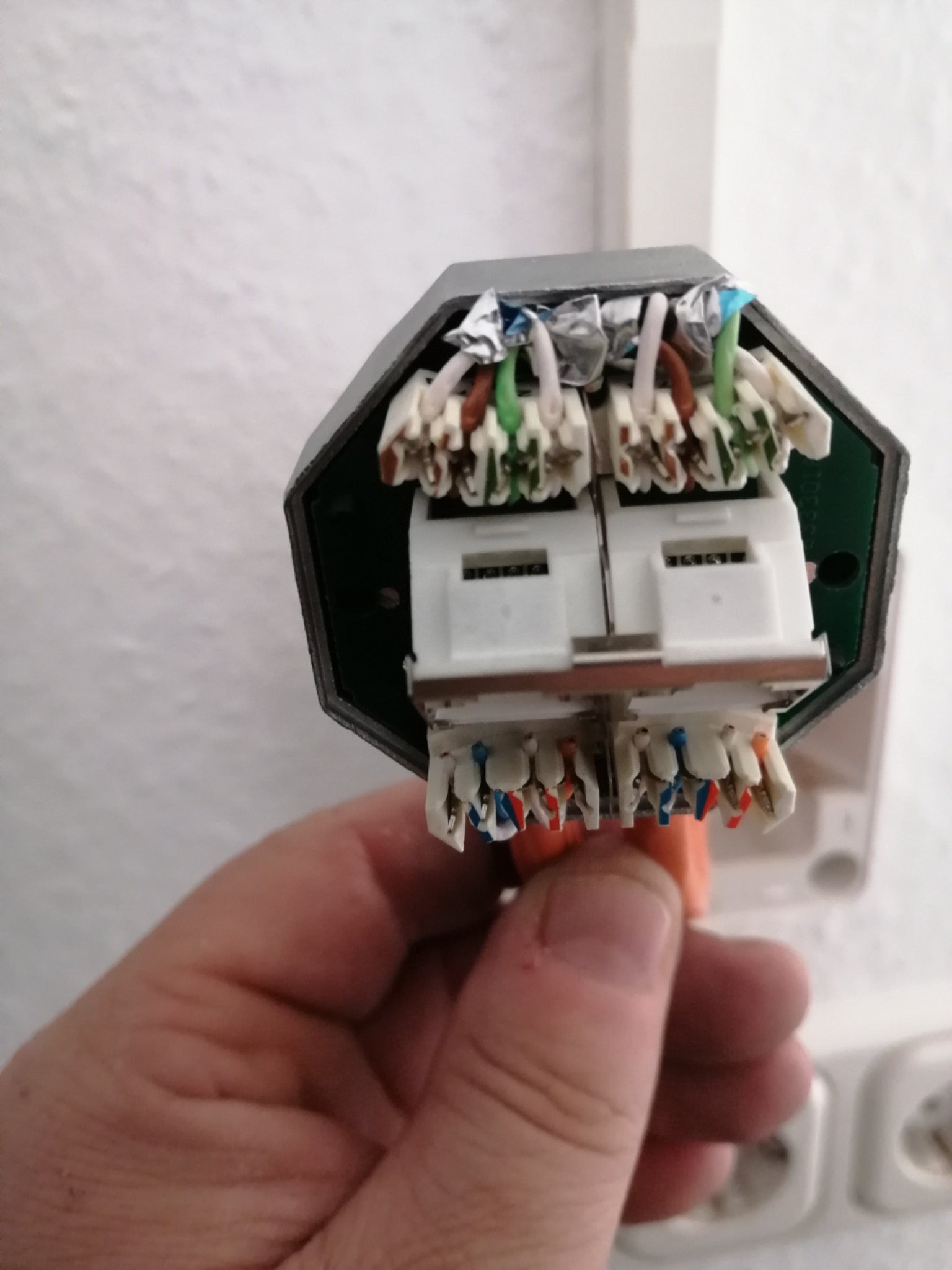 Szczegółowe ujęcie rozdzielnicy elektrycznej z podłączonymi przewodami o różnych kolorach, trzymanej w dłoni na tle białej ściany z gniazdkami.