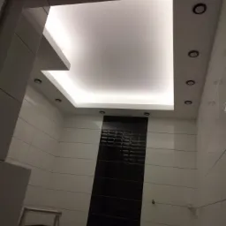 Nowoczesna łazienka z podświetlanym sufitem podwieszanym, czarno-białymi płytkami i drabinką w rogu.