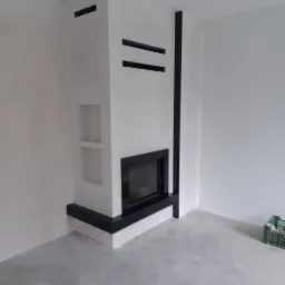 Nowoczesny kominek narożny z czarną obudową, w trakcie wykańczania ścian, widoczny surowy beton na podłodze i zielona skrzynka w rogu pomieszczenia.