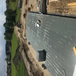 Widok z góry na płaski dach pokryty papą termozgrzewalną, z widocznym kominem wentylacyjnym i materiałami budowlanymi, na tle zieleni i błękitnego nieba.