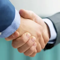 Uścisk dłoni dwóch mężczyzn w garniturach, niebieskim i szarym, na tle rozmytego, szklanego budynku.