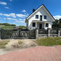 Nowoczesna, biała bryła domu z czarnymi akcentami, otoczona ogrodzeniem z metaloplastyki z motywem leśnym, osadzonym na kamiennych słupkach, w słoneczny dzień.