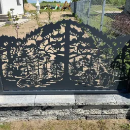 Metalowa brama wjazdowa z laserowo wycinanym wzorem leśnym przedstawiającym drzewa i wilki, zamontowana między kamiennymi słupkami.