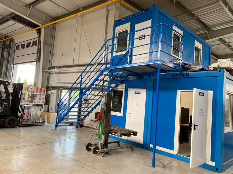 Dwupoziomowy, niebieski kontener biurowy z zewnętrznymi schodami metalowymi, umieszczony w hali produkcyjnej, widoczny wózek widłowy i regały z materiałami.